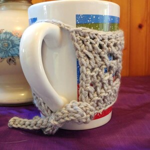 May include: A white ceramic mug with a gray crocheted cozy. The mug has a blue, green, and red design. The cozy is crocheted in a simple stitch pattern.