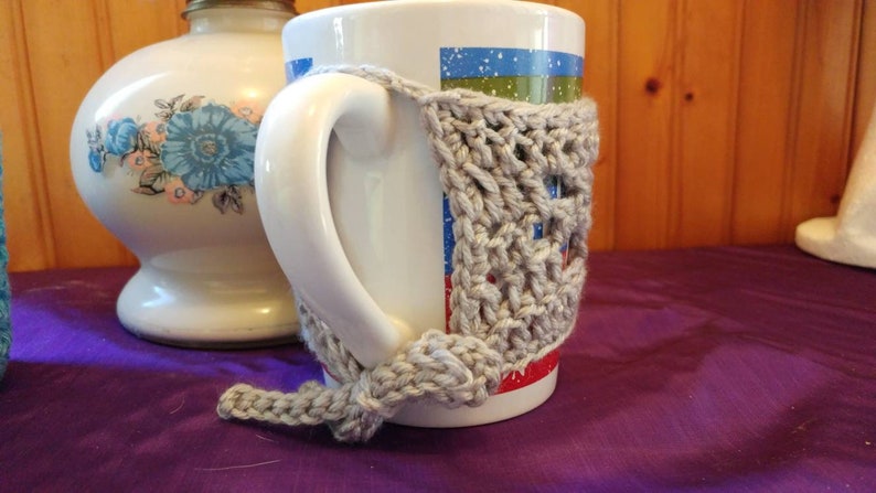 May include: A white ceramic mug with a gray crocheted cozy. The mug has a blue, green, and red design. The cozy is crocheted in a simple stitch pattern.