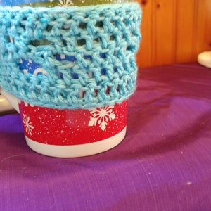 May include: A red and white mug with a blue crocheted cozy. The mug has a snowflake pattern.