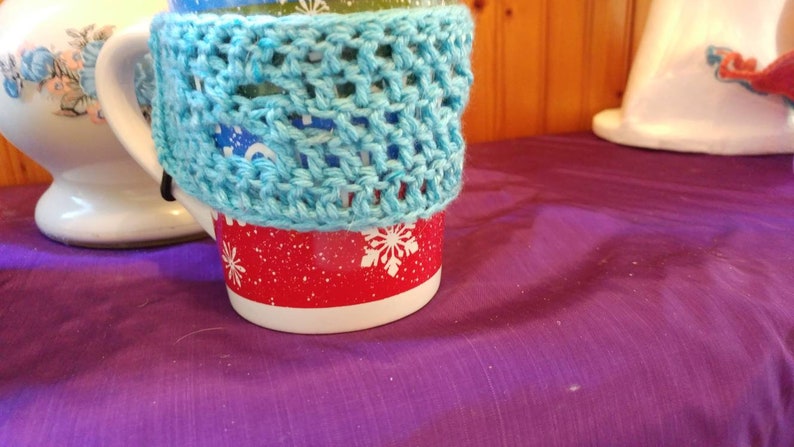 May include: A red and white mug with a blue crocheted cozy. The mug has a snowflake pattern.