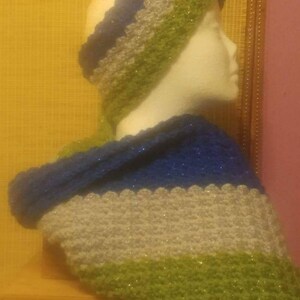 Puede incluir: Un conjunto de bufanda y diadema de ganchillo verde, plateado y azul. La bufanda est&aacute; drapeada sobre los hombros de un maniqu&iacute; y la diadema est&aacute; puesta en la cabeza del maniqu&iacute;.