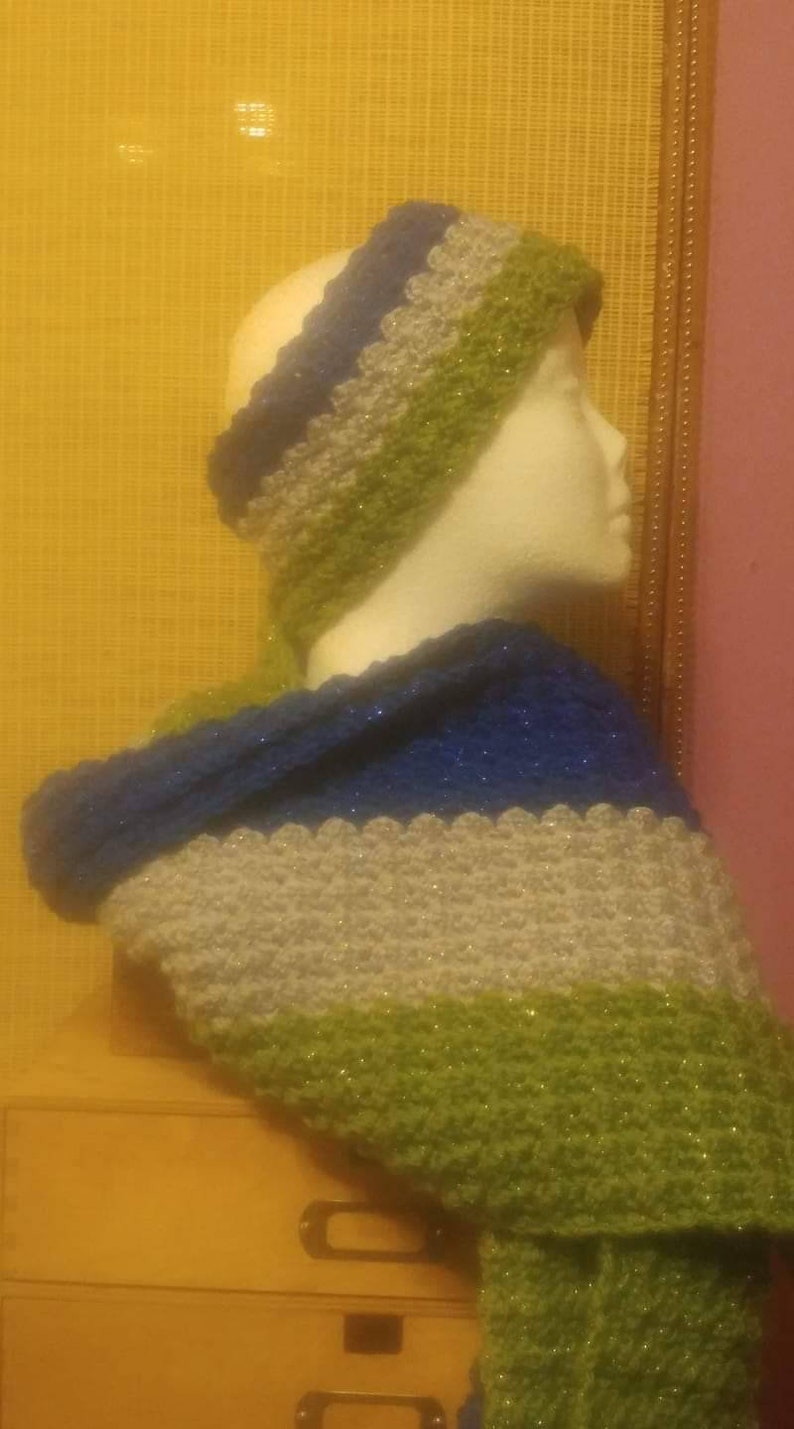 Puede incluir: Un conjunto de bufanda y diadema de ganchillo verde, plateado y azul. La bufanda est&aacute; drapeada sobre los hombros de un maniqu&iacute; y la diadema est&aacute; puesta en la cabeza del maniqu&iacute;.