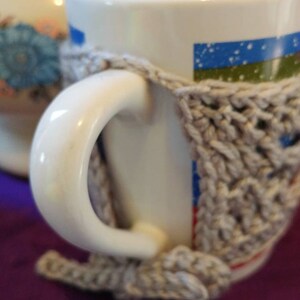 May include: A white ceramic mug with a blue and green striped design and a gray crocheted cozy. The mug is sitting on a purple surface.