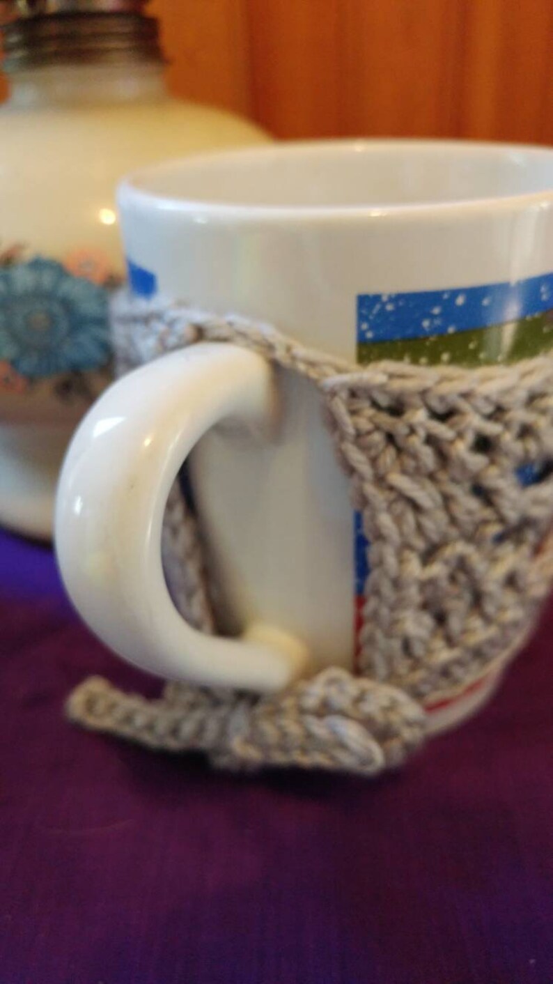 May include: A white ceramic mug with a blue and green striped design and a gray crocheted cozy. The mug is sitting on a purple surface.
