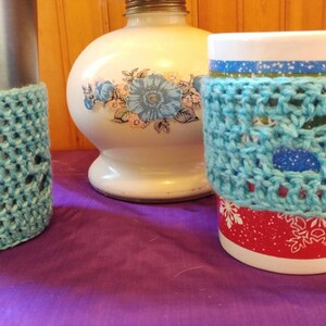 May include: Two crocheted cup cozies in a light blue color. The cozies are made with a lacy stitch pattern. One cozy is on a stainless steel travel mug and the other is on a white mug with a red and white snowflake design.