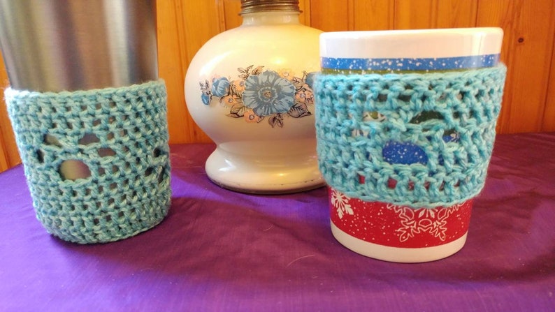 May include: Two crocheted cup cozies in a light blue color. The cozies are made with a lacy stitch pattern. One cozy is on a stainless steel travel mug and the other is on a white mug with a red and white snowflake design.