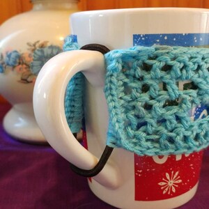 May include: White ceramic mug with a blue crocheted cozy around the handle. The mug has a red and white design with a snowflake on it.