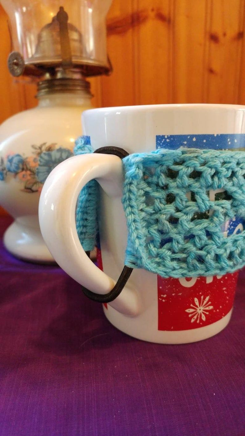 May include: White ceramic mug with a blue crocheted cozy around the handle. The mug has a red and white design with a snowflake on it.