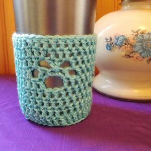May include: A light blue crocheted cup cozy with a paw print design. The cozy is on a stainless steel travel mug.