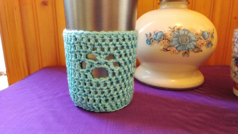 May include: A light blue crocheted cup cozy with a paw print design. The cozy is on a stainless steel travel mug.