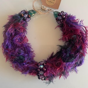 May include: A handmade necklace with a fluffy, textured design in shades of purple, pink, and black. The necklace features clusters of iridescent beads and green gemstone accents. A small tag reads "handmade with love."