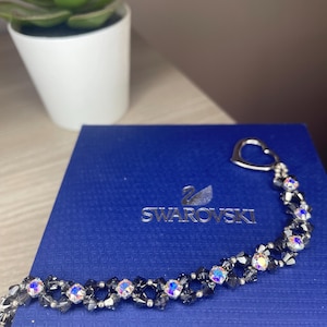 May include: A bracelet featuring dark gray faceted beads and iridescent crystals arranged in a repeating pattern, finished with a silver-toned heart-shaped clasp, resting on a blue Swarovski branded box with the company logo printed in silver.