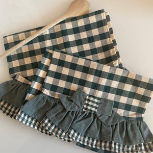 Op de afbeelding: Twee groene en witte geruite keukendoeken met geruite randen en een groene strik. De handdoeken zijn gevouwen en op elkaar gestapeld. Een houten lepel ligt op de handdoeken.