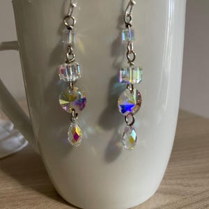 Può includere: Un paio di orecchini pendenti con cristalli trasparenti e iridescenti. Ogni orecchino presenta un gancio d'argento, una serie di piccole perline quadrate trasparenti, un cristallo rotondo più grande e un cristallo a forma di goccia. Gli orecchini sono esposti contro una tazza bianca.