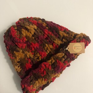 May include: A handmade, chunky knit beanie in a mix of brown, red, and gold colours. The hat has a folded brim and a small tag attached. The texture of the yarn is visible, creating a cosy and warm appearance.