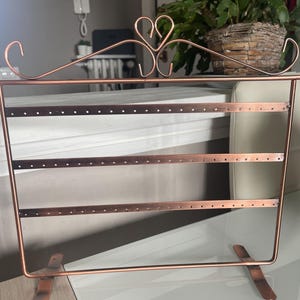 Pode incluir: Um expositor de joias de metal na cor cobre com três barras horizontais contendo orifícios espaçados uniformemente para pendurar brincos. O topo da estrutura é decorado com um design de arame central em formato de coração.
