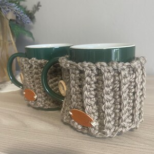 Op de afbeelding: Twee groene keramische mokken met gebreide beige hoezen. De hoezen zijn gemaakt van dik garen met een geribbelde textuur en zijn vastgezet met kleine bruine leren labels en houten knopen. De mokken staan op een licht houten oppervlak.