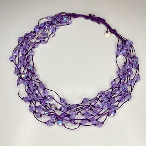 Puede incluir: Un collar de color morado de múltiples hilos con cuentas de color morado iridiscente. El collar está hecho de un cordón delgado y flexible y tiene un dije en forma de corazón.