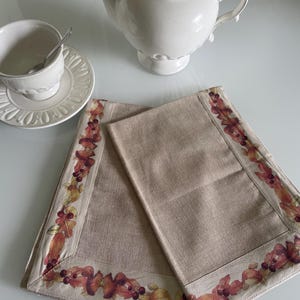 Può includere: Un set da tè in ceramica bianca con teiera, tazza e piattino con cucchiaino. Due tovagliette in lino beige con un bordo decorativo di foglie e bacche autunnali sono in primo piano. Il set da tè è su una superficie bianca.