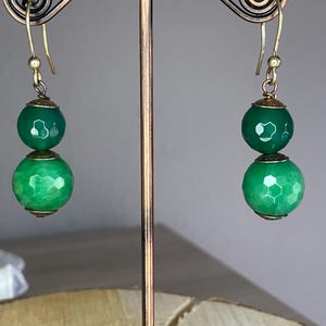 May include: A pair of dangle earrings featuring two stacked, faceted green beads on each side. The beads are separated and capped by small, antique-style bronze metal components. The earrings hang from simple bronze-toned metal ear wires.