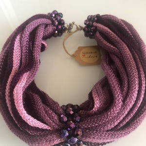 Peut inclure: Un collier en tissu tricoté violet et rose avec un groupe de perles violettes irisées au centre. Le collier est fait à la main et porte une étiquette indiquant "fait main avec amour".