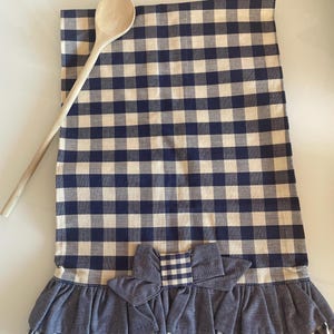 Może przedstawiać: Niebiesko-biały kratowany ręcznik kuchenny z niebieskim falbaniastym wykończeniem i niebieską kokardką. Ręcznik jest złożony, a na nim leży drewniana łyżka.