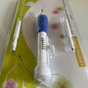 Puede incluir: Un juego de herramientas de aguja de perforación en un paquete de plástico transparente. El juego incluye una aguja de perforación blanca y azul, dos agujas adicionales y un enhebrador de alambre. El embalaje tiene un diseño floral verde y blanco y las palabras "Punch Needle".