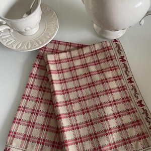 May include: Two folded beige and red plaid napkins with a decorative border featuring red bows and greenery. A white teacup with a spoon and a white teapot are also in the image.