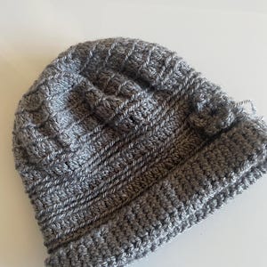 Peut inclure: Un bonnet en tricot gris fait à la main. Le bonnet est fait d'une laine douce et texturée et présente un bord côtelé. Le corps principal du bonnet a un motif de point texturé. Le bonnet est de couleur neutre.