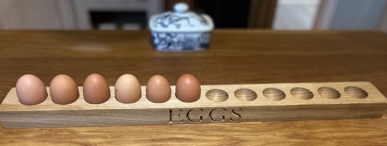May include: A wooden egg holder with the word "EGGS" engraved on the front. The holder has 10 slots for eggs, and 5 eggs are in the holder.