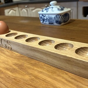 May include: A wooden egg holder with the word "EGGS" carved into the front. The holder has eight slots for eggs and is made of light brown wood.
