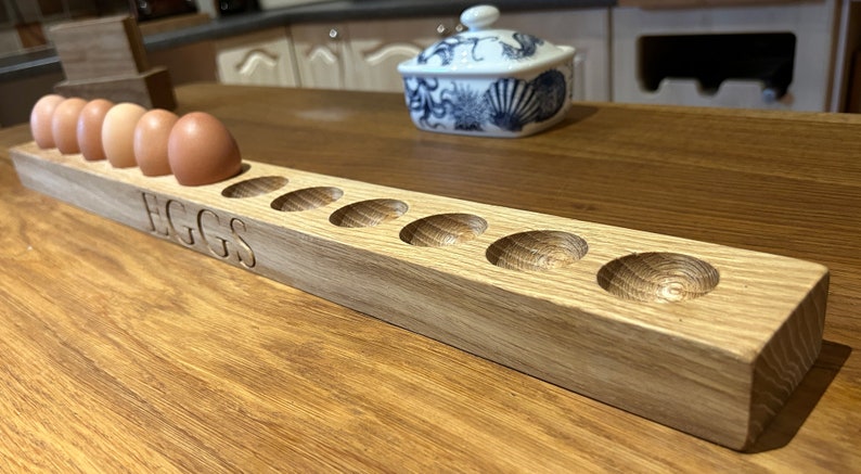 May include: A wooden egg holder with the word "EGGS" carved into the front. The holder has eight slots for eggs and is made of light brown wood.