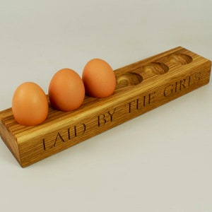 May include: Wooden egg holder with six slots, featuring the inscription "LAID BY THE GIRLS". Four brown eggs are placed in the holder.
