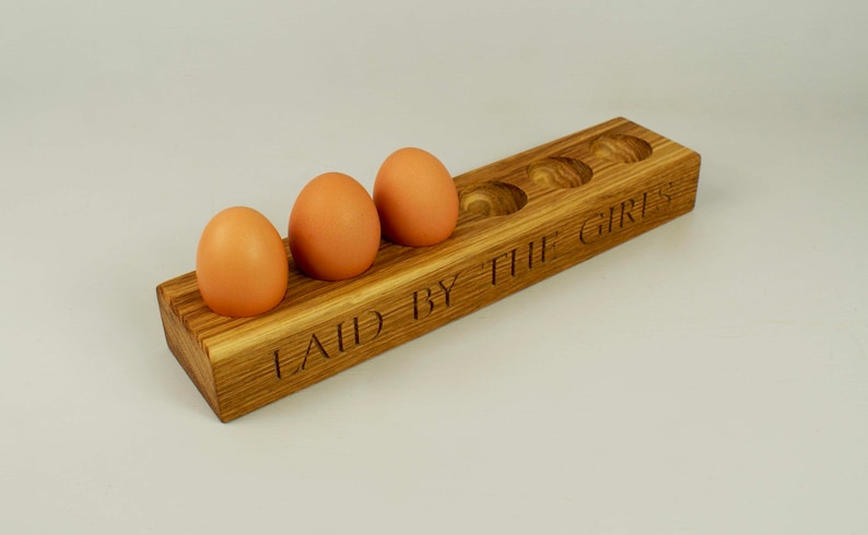 May include: Wooden egg holder with six slots, featuring the inscription "LAID BY THE GIRLS". Four brown eggs are placed in the holder.