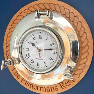 May include: A nautical-themed wall clock with a silver-colored porthole design. The clock face has Roman numerals and the brand name "Nauticalia". The clock is mounted on a wooden base with a rope-like border and the text "The Fishermans Rest".