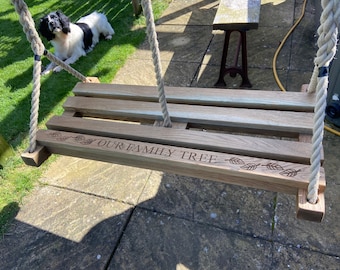 Op de afbeelding: Een houten schommel met touw dat van boven hangt. De schommel is gegraveerd met de woorden "Our Family Tree" en heeft bladontwerpen aan beide kanten van de tekst.