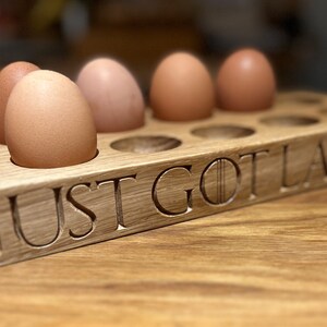 May include: A wooden egg holder with the text "JUST GOT LAID" carved into the front. Six brown eggs are sitting in the holder.