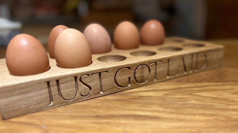 May include: A wooden egg holder with the text "JUST GOT LAID" carved into the front. Six brown eggs are sitting in the holder.