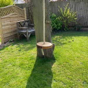 Könnte beinhalten: Ein hölzerner Schaukelsitz, der an einem Seil hängt, das an einem Baum in einem Garten befestigt ist. Der Schaukelsitz ist aus Naturholz gefertigt und hat eine Schlaufe aus Seil zum Aufhängen.