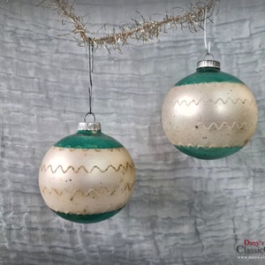 Könnte beinhalten: Zwei vintage Weihnachtskugeln aus Glas mit einem grün-weißen Farbschema. Die Kugeln haben ein weißes Band mit einem wellenförmigen goldenen Besatz um die Mitte.
