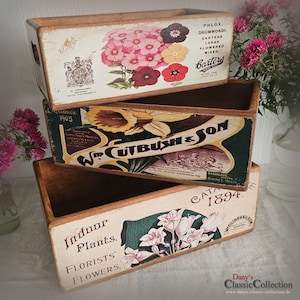 Könnte beinhalten: Drei rechteckige Holzpflanzkästen mit gealterten Oberflächen. Der obere Pflanzkasten zeigt ein Blumendesign mit Text, darunter "PHLOX. DRUMMONDI." Der mittlere Pflanzkasten hat ein Vintage-Etikett mit "W. CUTBUSH & SON." Der untere Pflanzkasten lautet "Indoor Plants."