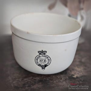May include: A white ceramic pudding boiler with a rounded shape and a wide rim. The front features a black emblem with the text "The Queen's Pudding Boiler No. 16 Patent". A classic kitchenware item.