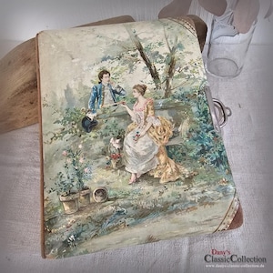 May include: Vintage book with a painted scene of a couple in a garden. The cover features a man in a blue jacket and a woman in a yellow dress. The scene includes a dog, flowers, and a bench. The book has a metal clasp and a brown border.