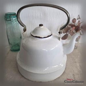 Könnte beinhalten: Eine weiße Emaille-Teekanne mit gebogenem Metallgriff und Ausguss. Die Teekanne hat einen abgerundeten Körper und einen Deckel mit einem kleinen Knopf. Ein Vintage-Glas und Trockenblumen sind im Hintergrund.
