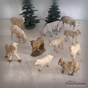 Peut inclure: Une collection de dix petites figurines de moutons dans diverses nuances de beige, crème et gris, accompagnées de deux arbres miniatures en pin vert foncé. Les moutons sont représentés debout ou en train de paître, avec des détails de laine texturés.