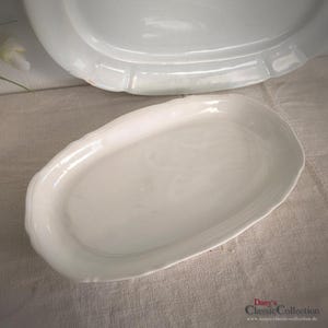 May include: A white, oval serving platter with a decorative, scalloped edge. The platter is made of ceramic and has a glossy finish. Another larger platter is partially visible in the background. The image is taken from a slightly elevated angle.