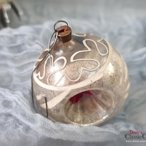 Puede incluir: Un adorno navideño vintage con acabado plateado y detalles decorativos blancos. El adorno tiene forma redonda con base acampanada y un lazo metálico para colgar. El adorno está sobre una tela azul claro.