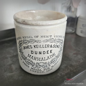 May include: A vintage white ceramic marmalade jar with black text and a decorative border. The jar reads "JAMES KEILLER & SONS DUNDEE MARMALADE" and includes text about awards. The jar is on a gray surface.