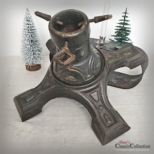 Könnte beinhalten: Antiker Weihnachtsbaumständer aus Bronze mit aufwendigen Details. Der Ständer hat einen kreisförmigen Sockel mit einer zentralen Aufnahme für den Baumstamm und verstellbaren Schrauben. Eine kleine weiße Kerze und zwei Miniatur-Weihnachtsbäume sind ebenfalls sichtbar.