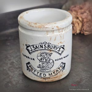 May include: Vintage white ceramic jar with a cylindrical form. The jar is marked with "J. Sainsbury's Potted Meats" in black script. The rim and exterior show signs of age and wear, enhancing its antique character.